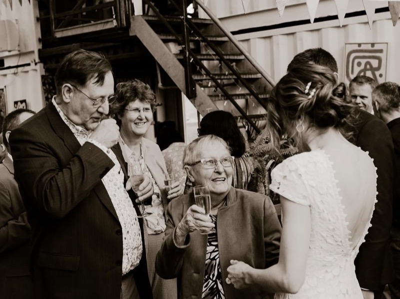 Oudere gast heft lachend het champagneglas naar de bruid, sepia